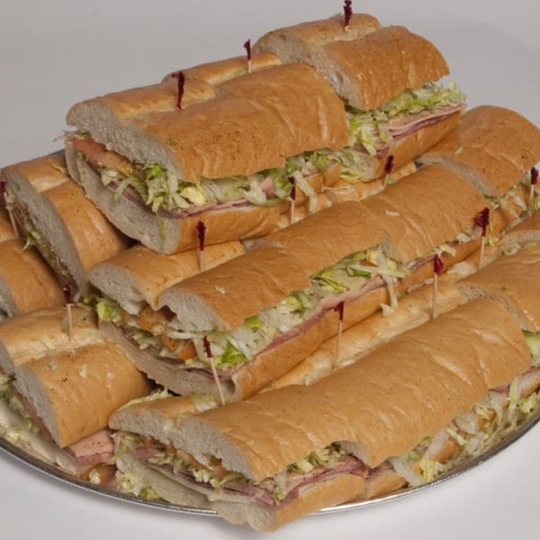 A catering tray filled with numerous long submarine sandwiches, cut into halves, revealing meat, cheese, and shredded lettuce.